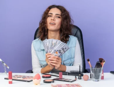 pleased-pretty-caucasian-woman-sitting-table-with-makeup-tools-holding-money